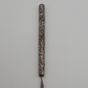 May include: A silver-toned metal stylus or writing tool featuring an intricate, embossed scrollwork pattern covering the entire cylindrical body. The tool has a pointed, diamond-shaped tip at one end and a flat, circular cap at the other end.