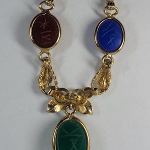 Puede incluir: Un collar dorado con tres piedras preciosas de forma ovalada: una roja, una azul y una verde. Cada piedra tiene un dise&ntilde;o grabado. El collar tiene un dise&ntilde;o decorativo de hojas y flores.