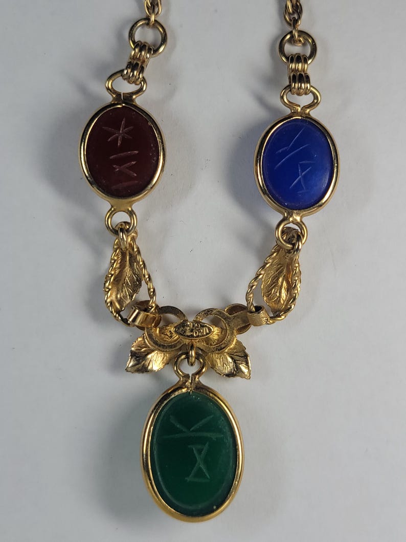 Puede incluir: Un collar dorado con tres piedras preciosas de forma ovalada: una roja, una azul y una verde. Cada piedra tiene un dise&ntilde;o grabado. El collar tiene un dise&ntilde;o decorativo de hojas y flores.