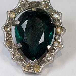 May include: Vintage silver-toned brooch featuring a large, pear-shaped, dark green gemstone at its center. The gemstone is surrounded by a decorative frame with smaller, clear and yellow gemstones. The brooch has a classic, elegant design.