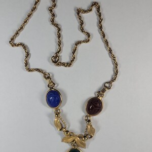 Puede incluir: Un collar de tono dorado con una cadena trenzada y tres colgantes ovalados estilo escarabajo en azul, rojo oscuro y verde. Los colgantes est&aacute;n unidos por eslabones de metal dorado y peque&ntilde;os detalles en forma de hoja.