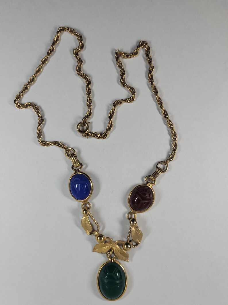 Puede incluir: Un collar de tono dorado con una cadena trenzada y tres colgantes ovalados estilo escarabajo en azul, rojo oscuro y verde. Los colgantes est&aacute;n unidos por eslabones de metal dorado y peque&ntilde;os detalles en forma de hoja.