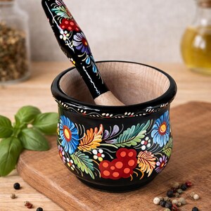 Może przedstawiać: Czarny zestaw moździerza i tłuczka z kwiatowym wzorem w kolorze niebieskim, czerwonym, żółtym i zielonym. Moździerz jest wykonany z drewna i ma pasujący tłuczek. Zestaw znajduje się na drewnianej desce do krojenia.