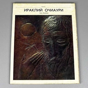 May include: A rectangular book cover featuring a dark, textured relief artwork of a bearded face and a hand. The text at the top reads "ИРАКЛИЙ ОЧИАУРИ" and "Кити Мачабели" in black Cyrillic lettering against a cream-colored border background.