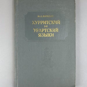 May include: A vintage book with a gray cover. The title, in gold lettering, reads "HURRITSKY and URARTIAN LANGUAGES." The author's name, "M.A. KHACHIKYAN," is above the title. A small embossed design is visible on the lower right.