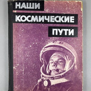 Op de afbeelding: Vintage boekomslag met de titel "НАШИ КОСМИЧЕСКИЕ ПУТИ" in witte letters op een zwarte achtergrond. De omslag toont een portret van een kosmonaut in een ruimtehelm tegen een paarse sterrenachtergrond.