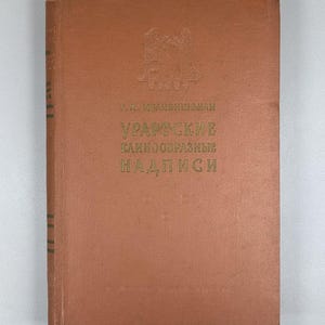 May include: A vintage, hardcover book with a brown cover. The title, in green text, reads "УРАРТСКИЕ КЛИНООБРАЗНЫЕ НАДПИСИ". The book has a faded appearance, suggesting age. The cover features an embossed illustration and the author's name.