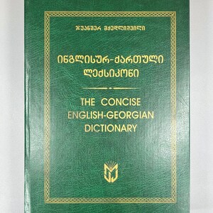 May include: A green book with gold lettering and a gold border. The text on the cover reads "THE CONCISE ENGLISH-GEORGIAN DICTIONARY" and includes text in Georgian script. A gold emblem is at the bottom.