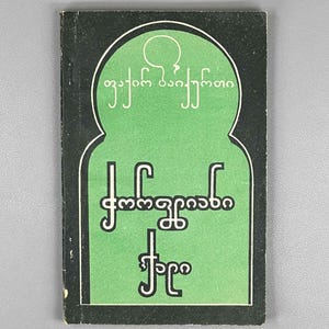 Op de afbeelding: Een rechthoekig boek met een zwarte kaft en een centraal groen boogvormig ontwerp. Het groene gedeelte bevat witte tekst in een niet-Latijns schrift, waaronder de tekens "ფაირ ბაიქურთი" bovenaan en "ქორწინებიანი ქალი" daaronder.