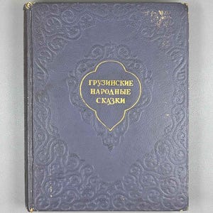 Pode incluir: Um livro de capa dura com uma capa texturizada azul-escura apresentando um padrão floral em relevo. O centro exibe um cartucho com contorno dourado contendo o texto cirílico "ГРУЗИНСКИЕ НАРОДНЫЕ СКАЗКИ" em letras douradas.