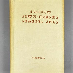 May include: A hardcover book with a light beige cover. The title is in red Georgian script. The book's title is "Georgian Dialect Dictionary". The word "Ganatleba" is printed at the bottom of the book.