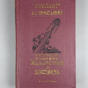 May include: A maroon-colored book with gold lettering and illustrations. The cover features text in a foreign language and a black illustration of a stylized object. The book appears to be vintage or antique.