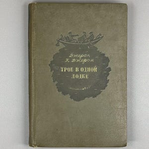 May include: An old book with a muted olive-green cover. The cover displays a boat illustration with figures and Cyrillic text. The title is "Three Men in a Boat" by Jerome K. Jerome.
