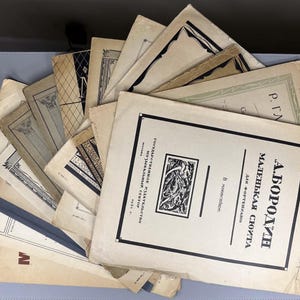 May include: A collection of vintage sheet music with aged, off-white covers. The covers feature various designs, including geometric patterns and text in a foreign language. The sheet music is arranged in a fan-like display, suggesting a collection of musical scores.