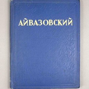 Op de afbeelding: Een blauw boek met de titel "АЙВАЗОВСКИЙ" in witte letters. Het boek heeft een getextureerde omslag en een rechthoekige vorm. De titel is geschreven in Cyrillisch schrift.