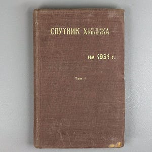 May include: A vintage brown book with gold lettering. The title reads "Спутник Химика" above "на 1931 г." and "Том 1". The book has a worn appearance, suggesting age and use. The background is a light grey surface.
