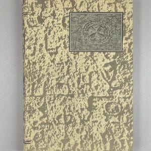 Op de afbeelding: Een vintage boek met een getextureerde, beige en grijze omslag. De omslag heeft een herhalend patroon en een rechthoekige inzet met een kruisontwerp. De rug heeft twee donkergrijze lijnen.