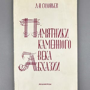 May include: A vintage book with a cream-colored cover. The title, in maroon Cyrillic script, reads "Monuments of the Stone Age of Abkhazia." The author's name is at the top. The publisher's name is at the bottom.
