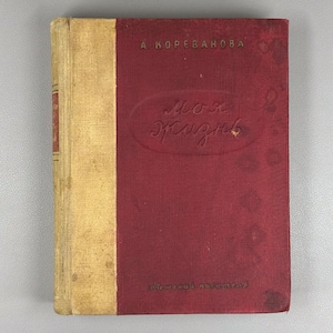 May include: A vintage book with a red cover and gold spine. The cover features embossed text in a cursive font, with the title "My Life" visible. The author's name, "A. Korevanova," is printed at the top. The book's edges show signs of age.