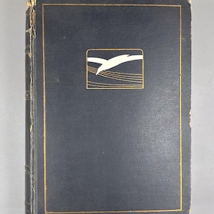 May include: A vintage, navy blue book with a gold border and a gold-framed emblem featuring a white bird in flight. The book's cover shows signs of wear, with a slightly distressed appearance.