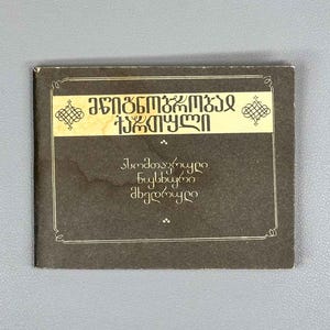 Op de afbeelding: Een klein, donkerbruin boek met gouden tekst en decoratieve elementen. De omslag bevat tekst in een uniek lettertype, met een gouden banner bovenaan. Het boek staat tegen een grijze achtergrond.