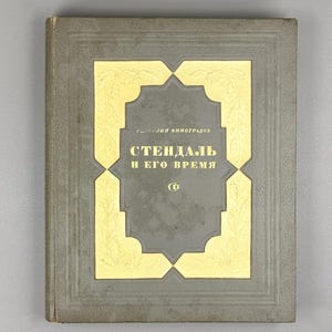 Pode incluir: Um livro vintage com capa cinza e detalhes dourados. O título, em russo, é "Stendhal e seu tempo". A capa apresenta uma moldura decorativa dourada em torno do título. O livro tem uma aparência desgastada.