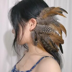 May include: A decorative hair accessory featuring a cluster of brown, gray, and white spotted feathers. The feathers are arranged to frame the ear, creating a unique and bohemian look. The accessory is designed to be worn in the hair.
