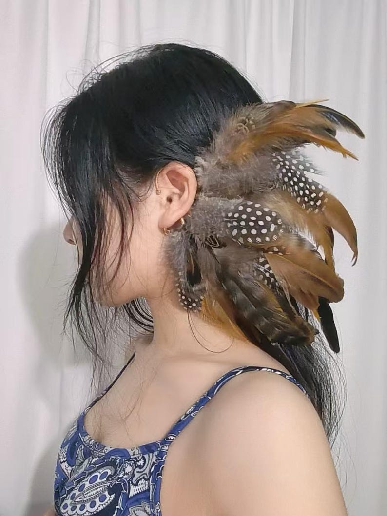 May include: A decorative hair accessory featuring a cluster of brown, gray, and white spotted feathers. The feathers are arranged to frame the ear, creating a unique and bohemian look. The accessory is designed to be worn in the hair.