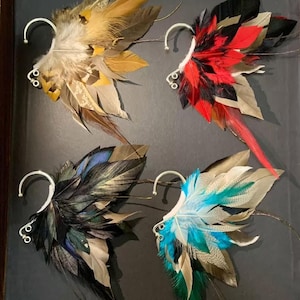 May include: Four feather ear cuffs in a display. Each cuff features a unique color scheme: tan and yellow, red and black, black and blue, and blue and green. The cuffs have a silver-colored ear wire.