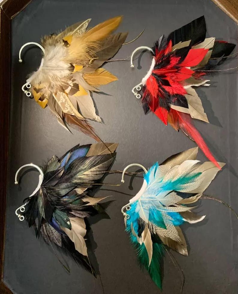 May include: Four feather ear cuffs in a display. Each cuff features a unique color scheme: tan and yellow, red and black, black and blue, and blue and green. The cuffs have a silver-colored ear wire.