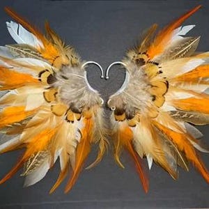 May include: Ear cuffs with a wing design made of orange, white, and brown feathers. The ear cuffs have a silver-colored metal heart-shaped frame. The feathers are arranged to resemble wings.