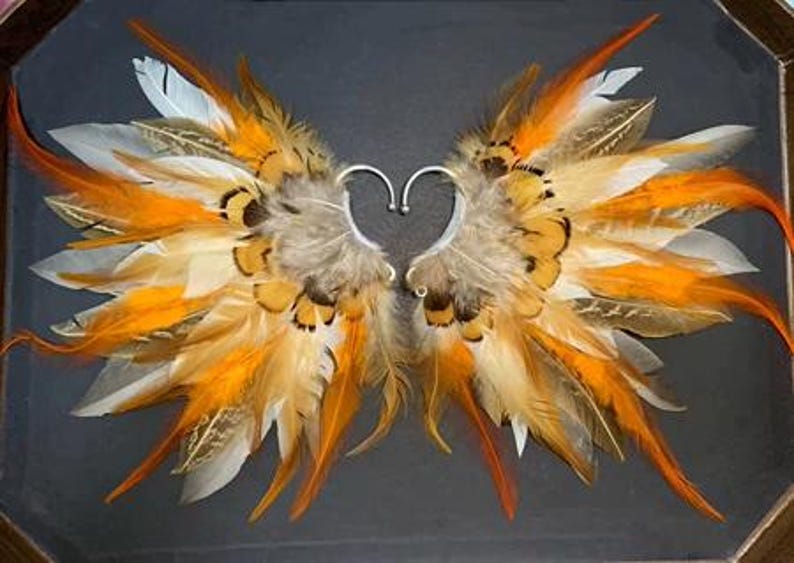 May include: Ear cuffs with a wing design made of orange, white, and brown feathers. The ear cuffs have a silver-colored metal heart-shaped frame. The feathers are arranged to resemble wings.