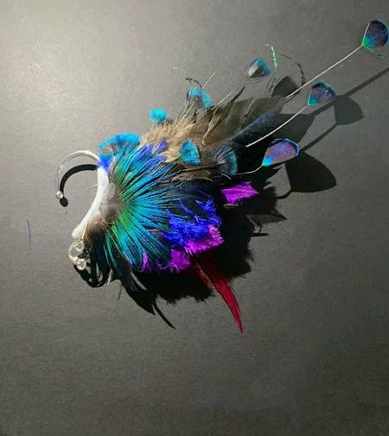 May include: A vibrant ear cuff featuring a peacock feather design. The cuff showcases a spectrum of colors, including teal, blue, purple, and red, with a silver-tone metal ear wrap. The feathers are arranged to create a striking, artistic accessory.