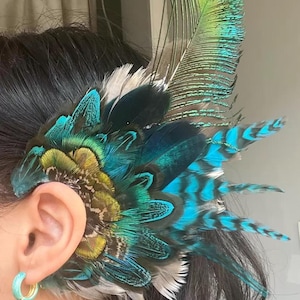 Puede incluir: Un brazalete de plumas vibrantes con una mezcla de plumas turquesas, verde azulado y amarillo verdoso. El accesorio est&aacute; dise&ntilde;ado para usarse en la oreja, con un grupo de plumas que se abren en abanico. Las plumas tienen una apariencia natural y texturizada.