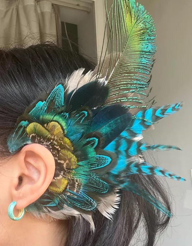 Puede incluir: Un brazalete de plumas vibrantes con una mezcla de plumas turquesas, verde azulado y amarillo verdoso. El accesorio est&aacute; dise&ntilde;ado para usarse en la oreja, con un grupo de plumas que se abren en abanico. Las plumas tienen una apariencia natural y texturizada.