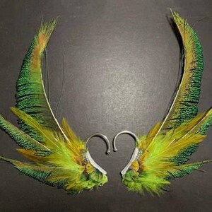 May include: A pair of handmade feather earrings. The earrings feature vibrant green, yellow, and brown feathers attached to silver-colored ear wires. The feathers are arranged in a wing-like shape, creating a unique and eye-catching accessory.