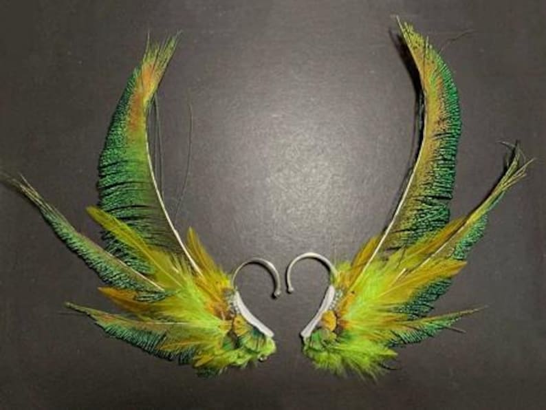 May include: A pair of handmade feather earrings. The earrings feature vibrant green, yellow, and brown feathers attached to silver-colored ear wires. The feathers are arranged in a wing-like shape, creating a unique and eye-catching accessory.