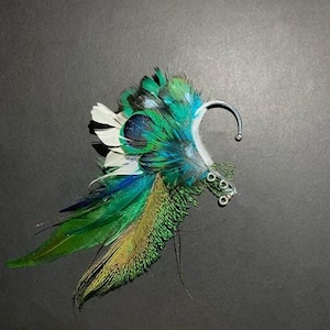 May include: A decorative ear cuff featuring vibrant green, blue, and white feathers. The feathers are arranged to create a wing-like design, with a silver-toned metal ear hook. The cuff is designed to be worn on the ear.