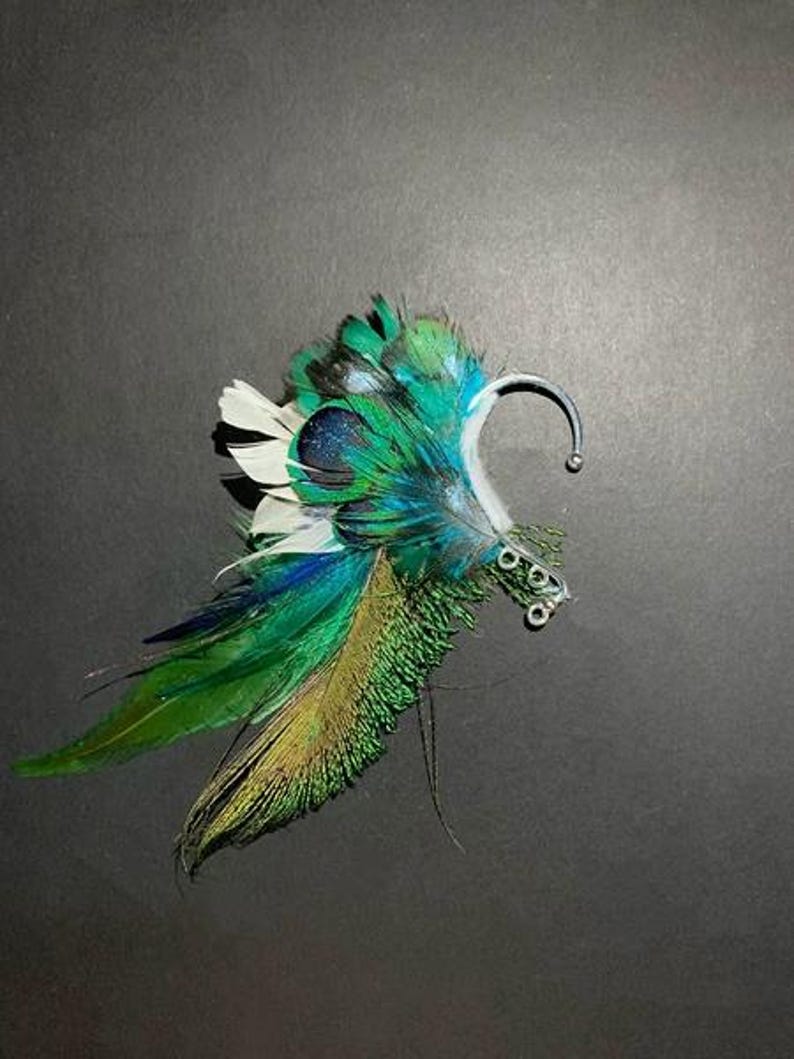 May include: A decorative ear cuff featuring vibrant green, blue, and white feathers. The feathers are arranged to create a wing-like design, with a silver-toned metal ear hook. The cuff is designed to be worn on the ear.