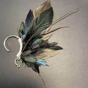 May include: An ear cuff featuring a cluster of feathers in shades of black, teal, brown, and blue. The feathers are attached to a silver-colored metal ear cuff with a curved design. The feathers are arranged to create a wing-like appearance.