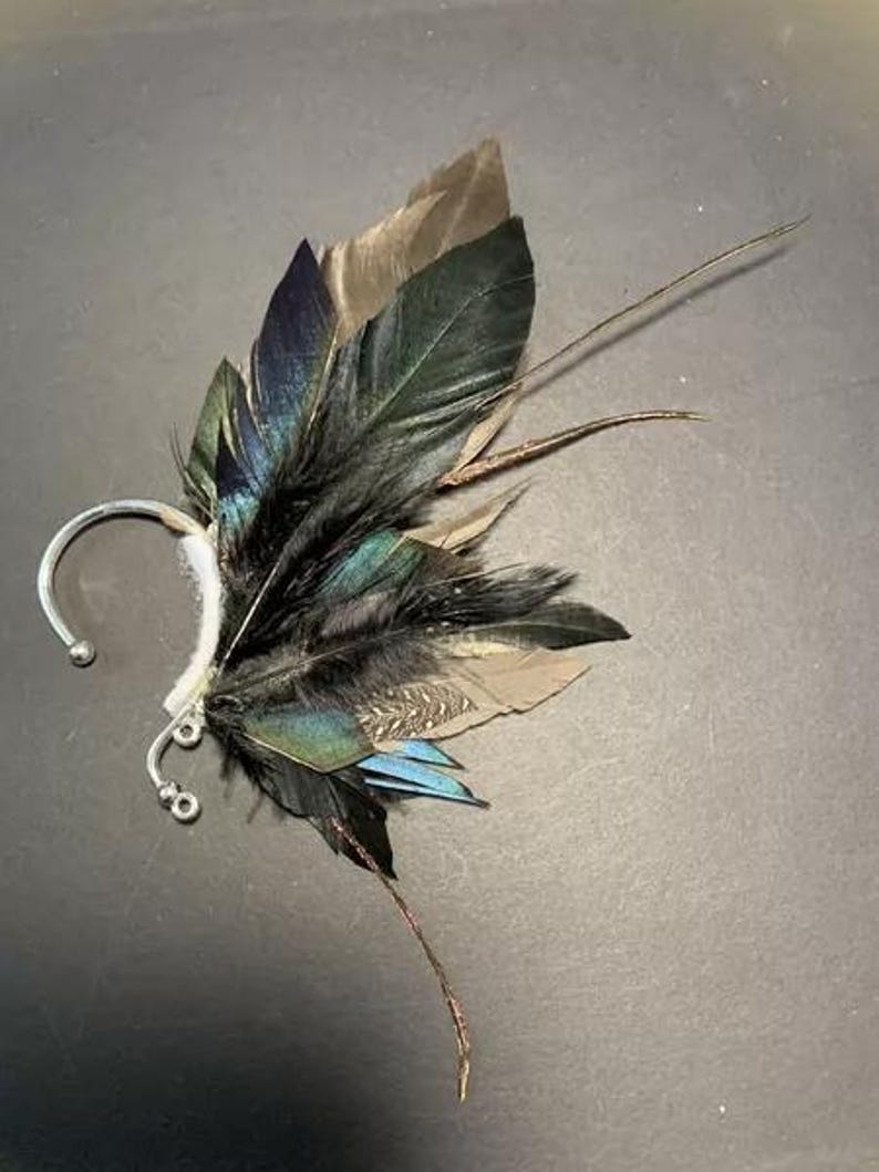 May include: An ear cuff featuring a cluster of feathers in shades of black, teal, brown, and blue. The feathers are attached to a silver-colored metal ear cuff with a curved design. The feathers are arranged to create a wing-like appearance.