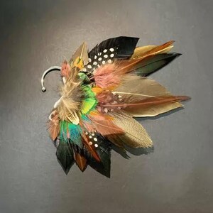May include: A colorful feather ear cuff featuring a variety of feathers in shades of brown, black, green, and pink. The ear cuff has a silver-colored hook for attachment. The feathers are arranged in a layered, fan-like design.