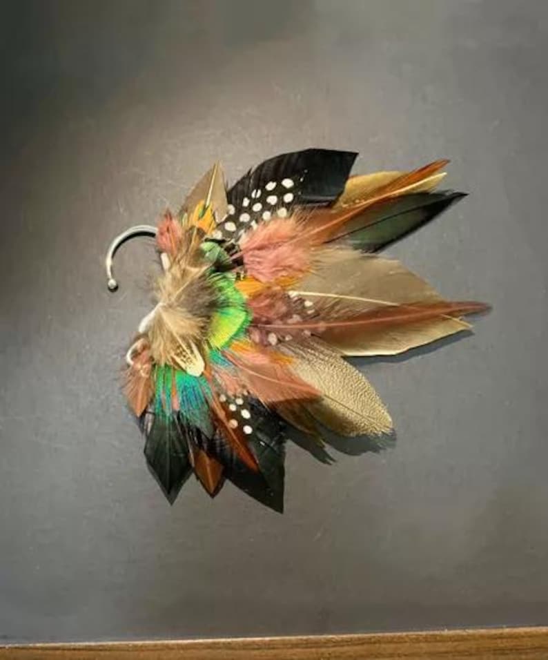 May include: A colorful feather ear cuff featuring a variety of feathers in shades of brown, black, green, and pink. The ear cuff has a silver-colored hook for attachment. The feathers are arranged in a layered, fan-like design.
