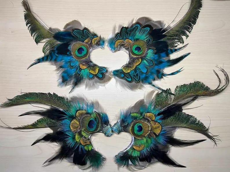 Puede incluir: Cuatro pares de m&aacute;scaras de ojos de plumas de pavo real. Cada m&aacute;scara presenta un dise&ntilde;o central de ojo con plumas azules, verdes y doradas. Las m&aacute;scaras est&aacute;n dispuestas en pares, con los ojos enfrentados.