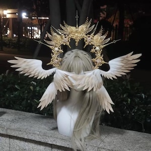 May include: A gold-toned halo headpiece featuring intricate filigree metalwork, star-shaped accents, and small white pearls. Attached to the headpiece are multiple sets of white feathered wings that extend outward from the sides and back.
