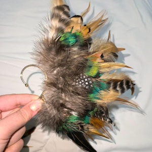 May include: An ear cuff featuring a cluster of feathers in shades of brown, green, and black. The feathers are attached to a gold-colored metal ear hook. The design includes a variety of feather textures and patterns, creating a unique accessory.
