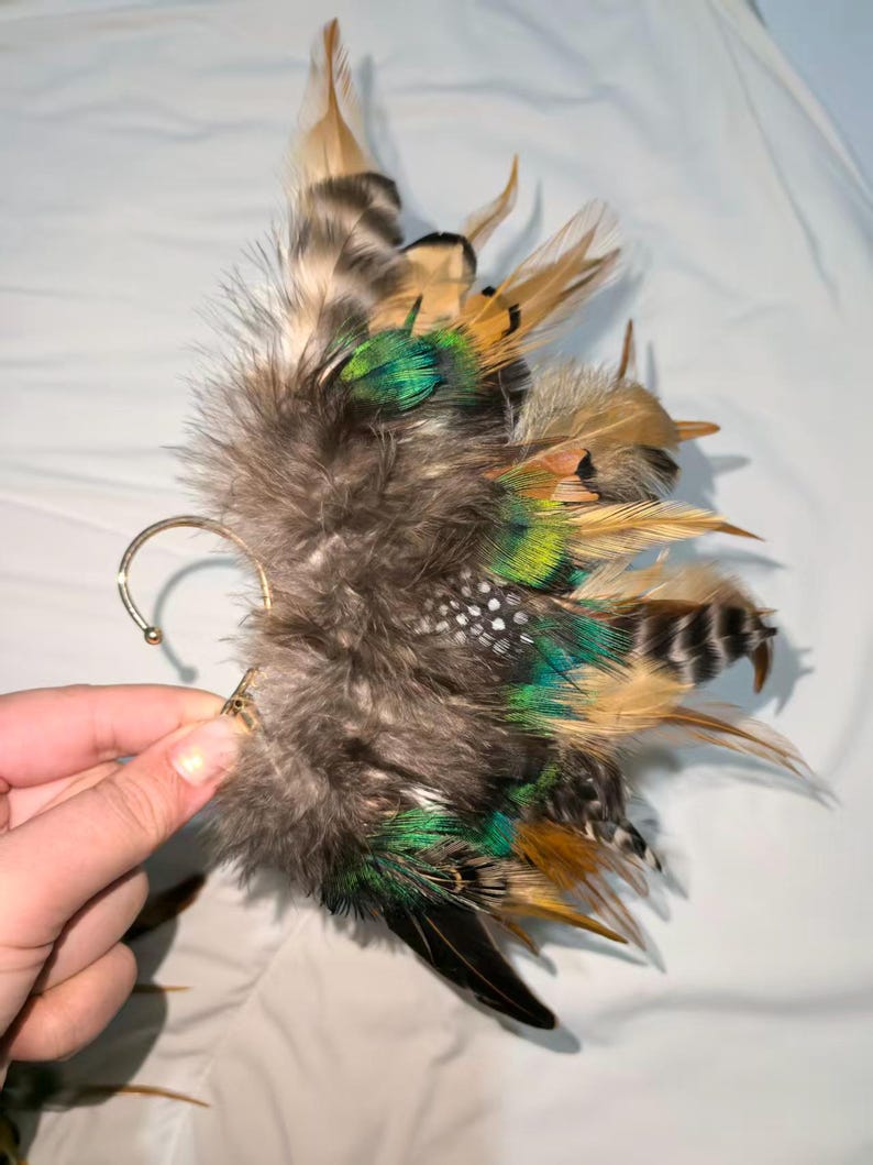 May include: An ear cuff featuring a cluster of feathers in shades of brown, green, and black. The feathers are attached to a gold-colored metal ear hook. The design includes a variety of feather textures and patterns, creating a unique accessory.