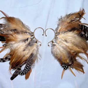 May include: A pair of ear cuffs featuring a feather design. The feathers are in shades of brown, tan, and black, with a metallic silver ear wire. The ear cuffs are designed to be worn on the ear, creating a unique and eye-catching accessory.