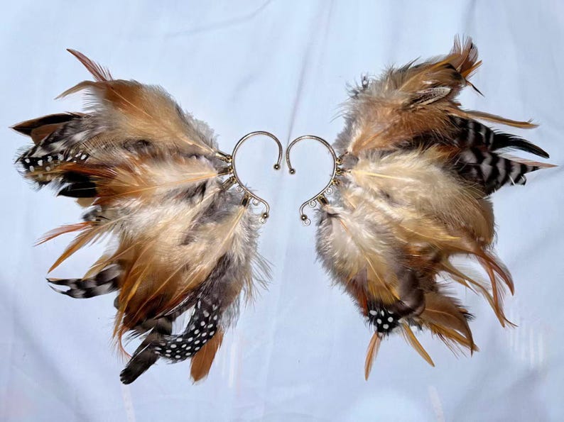 May include: A pair of ear cuffs featuring a feather design. The feathers are in shades of brown, tan, and black, with a metallic silver ear wire. The ear cuffs are designed to be worn on the ear, creating a unique and eye-catching accessory.