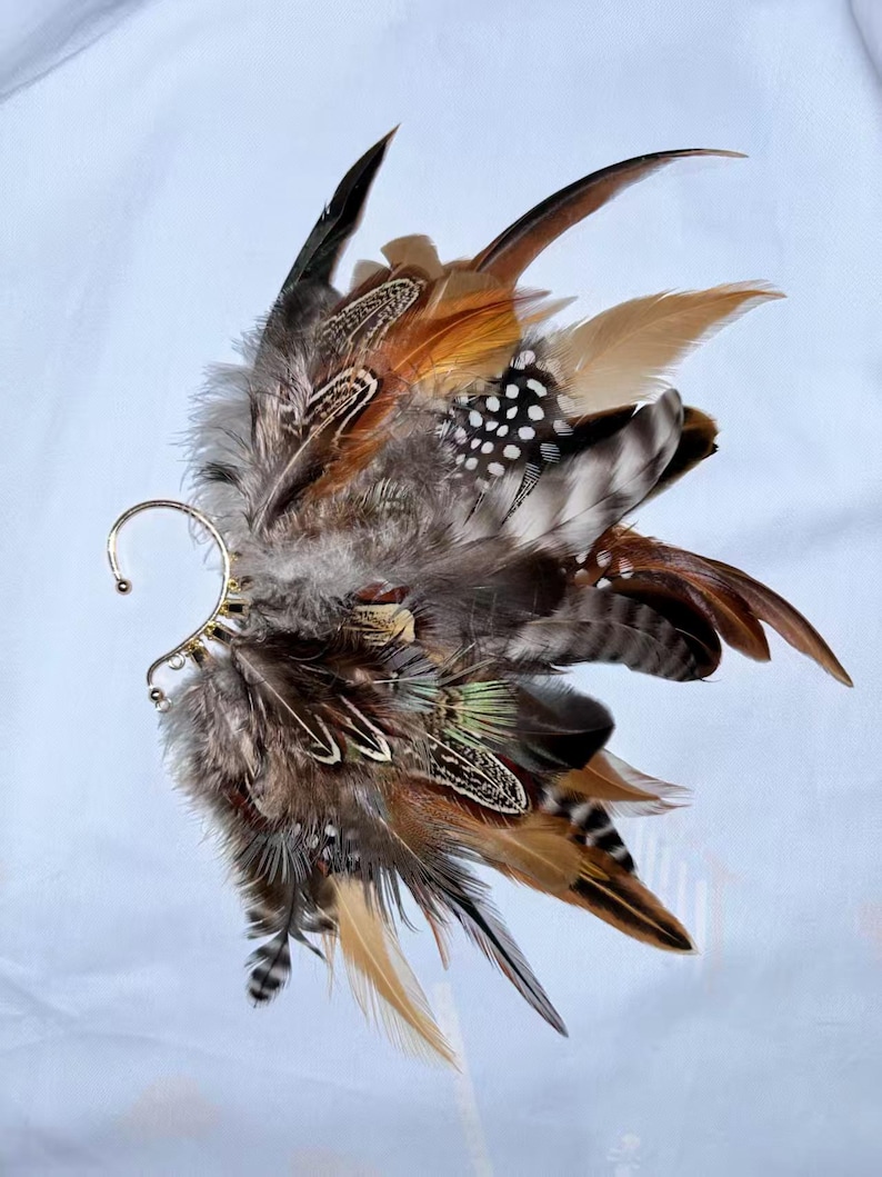 May include: An ear cuff featuring a cluster of feathers in shades of brown, tan, black, and white. The feathers are arranged in a layered design, with a gold-colored metal ear loop. The feathers have various patterns, including spots and stripes.